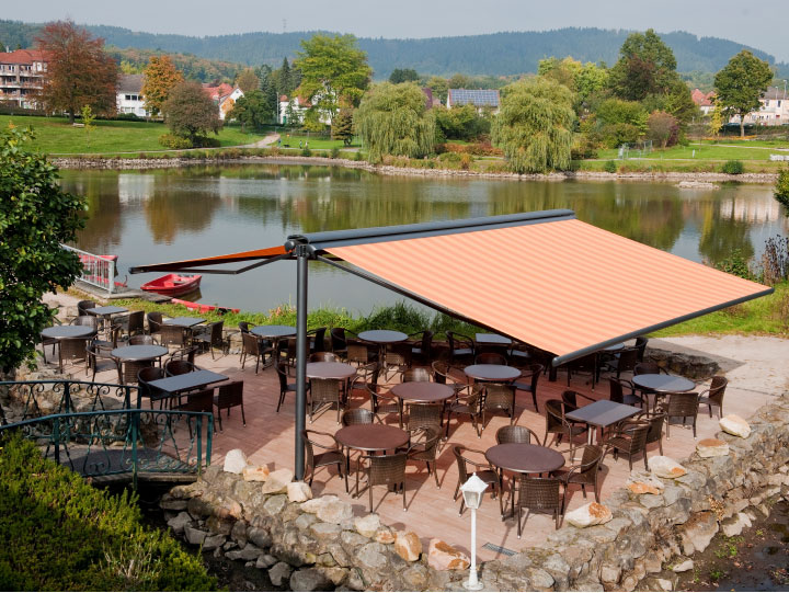 Image of syncra frame system fitted with cassetted folding-arm awnings at the seating area of a  lakeside restaurant.>
